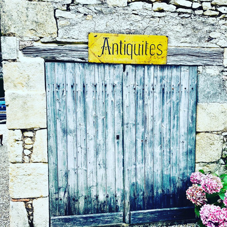 A weathered yellow tin sign hand-lettered ANTIQUITÉS in black, mounted on a rough oak beam above closed double doors of weathered grey planks set in a stone wall, with pink hydrangea blooming in the bottom right corner