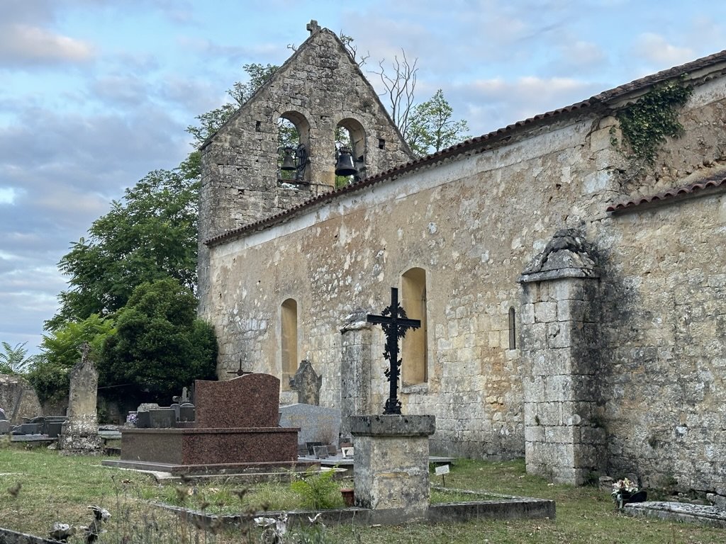 The Romanesque church of Saint-Martin near Bannes — a stone bell-gable with two iron bells, weathered limestone walls, a wrought-iron crucifix on a stone plinth, and a small graveyard with worn headstones in the foreground