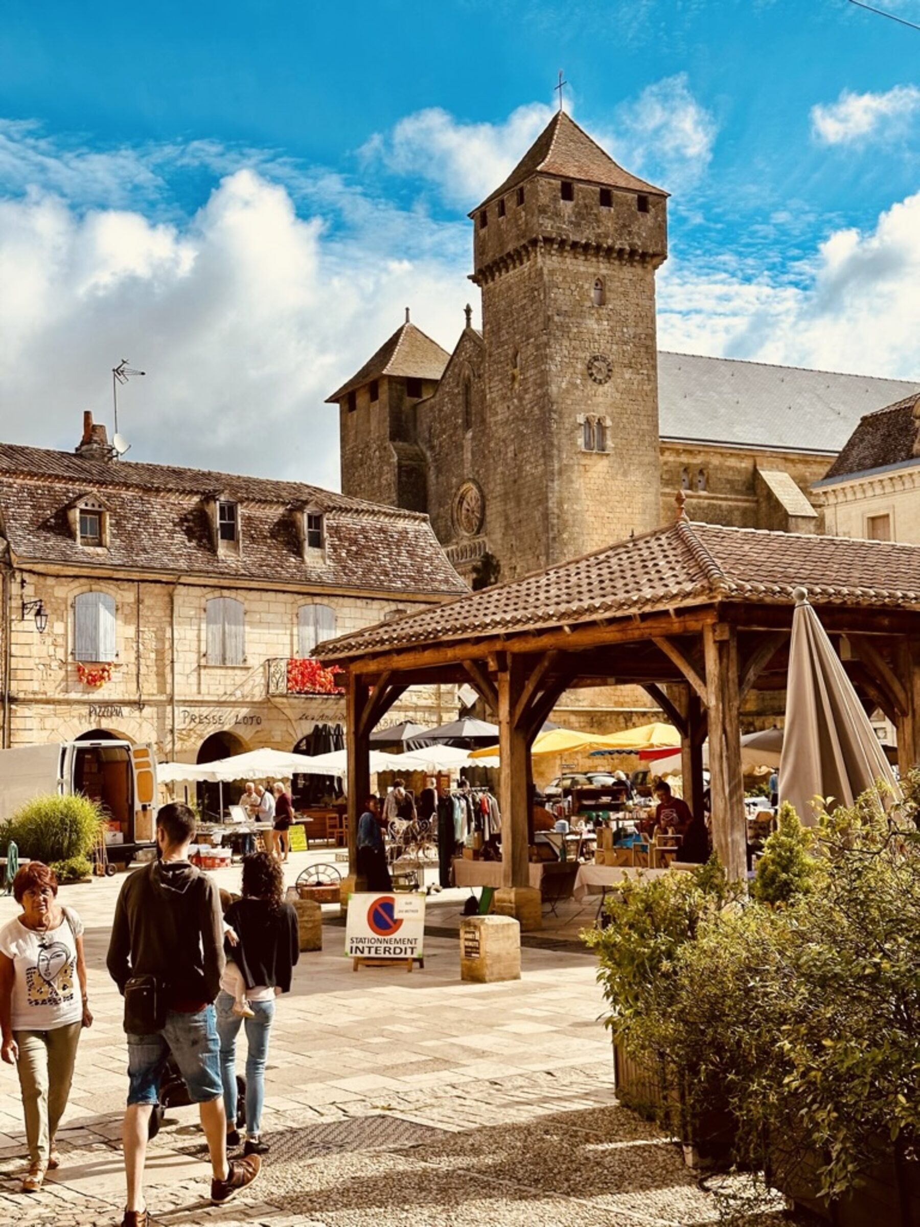Beaumont-du-Périgord bastide square on market day — the medieval fortified église Saint-Laurent with its twin defensive towers rising above warm-stone houses, the 13th-century covered wooden halle in the foreground with market stalls underneath, shoppers walking across the cobbled square under blue sky