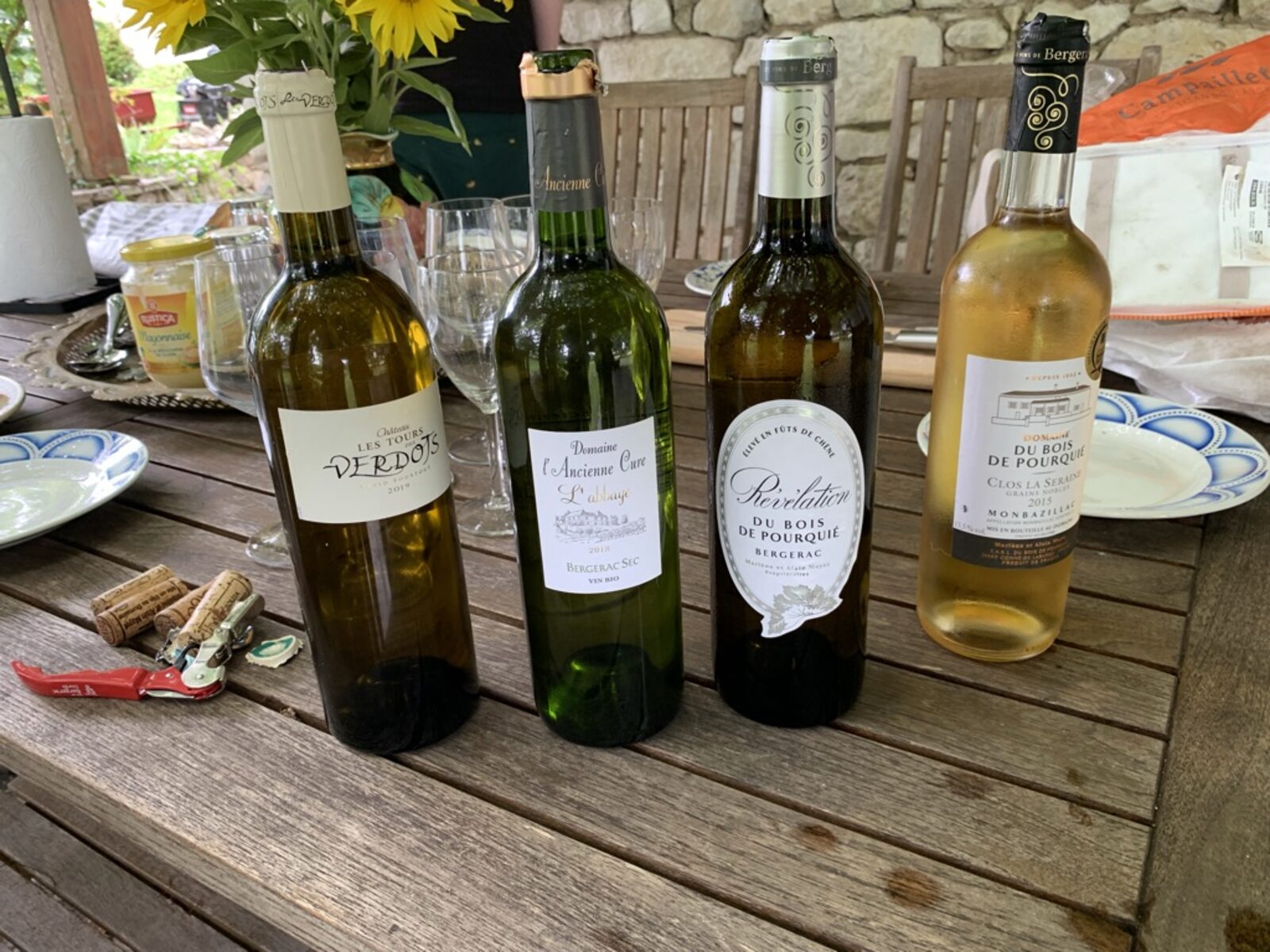 Four Bergerac bottles lined up on the long wooden table under the pergola — Château Les Tours des Verdots, Domaine de l'Ancienne Cure L'Abbaye (Bergerac Sec), Révélation Du Bois de Pourquié (Bergerac), and a Monbazillac from Clos La Seraine — corks pulled, sunflowers at the back, plates set