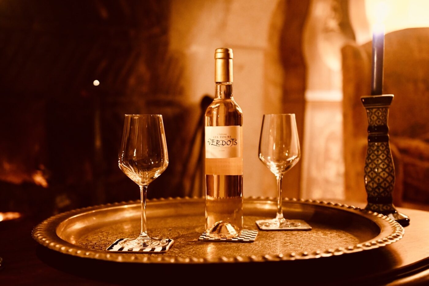 A small bottle of Château Les Tours des Verdots dessert wine on a Moroccan brass tray, two empty glasses, a candle, firelight warming the whole scene
