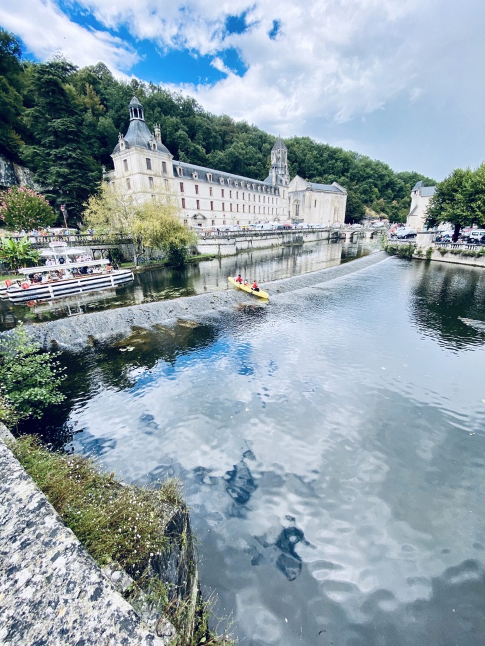 The Brantôme abbey seen from across the Dronne — a tall white Romanesque bell tower rising above limestone cliffs, the abbey buildings stretching along the riverbank, a pleasure boat moored below, a yellow canoe on the water