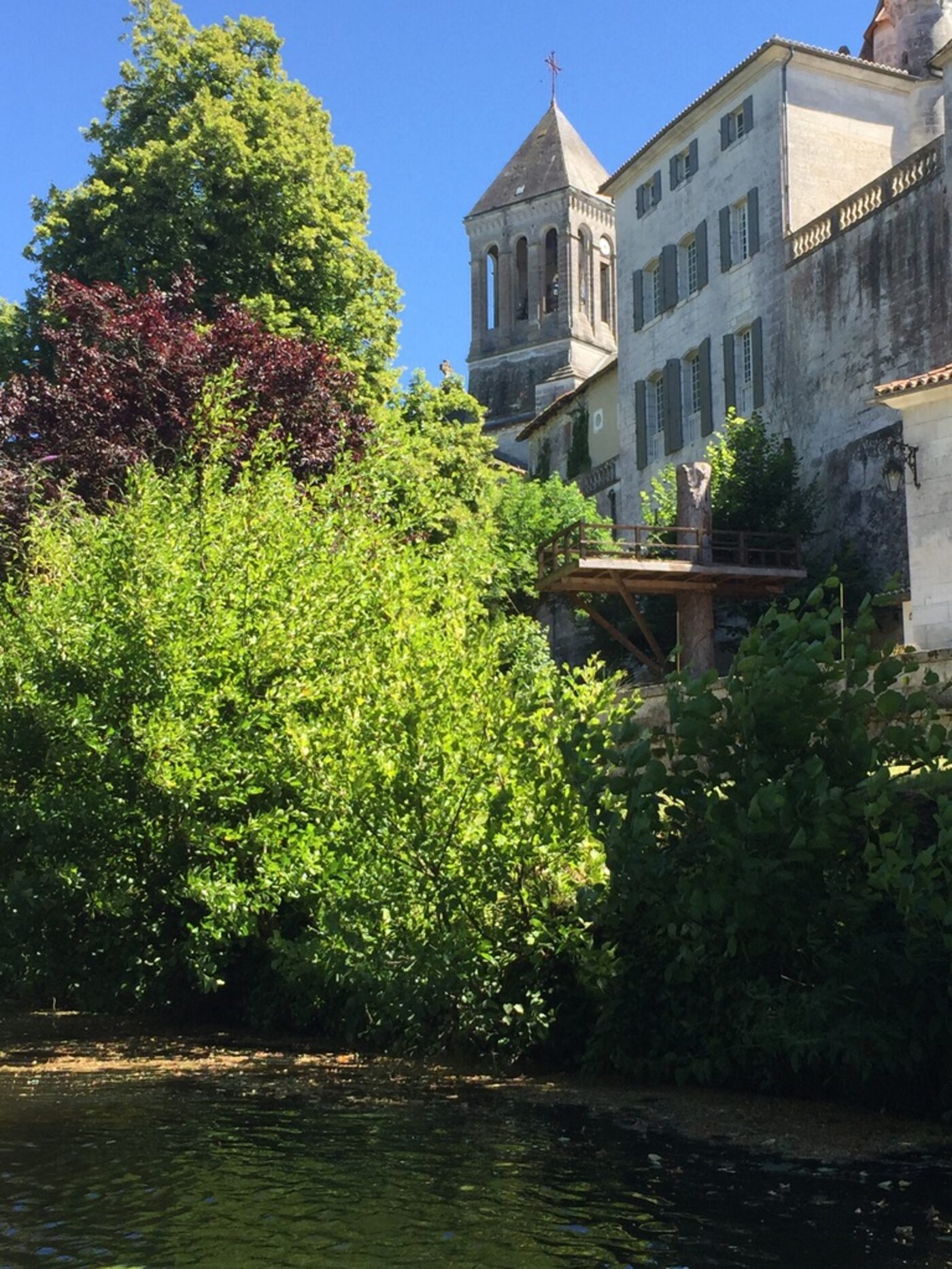 A glimpse of the Brantôme abbey bell tower rising through dense green trees on the edge of the Dronne river, a small wooden fishing platform jutting out over the water, summer foliage everywhere