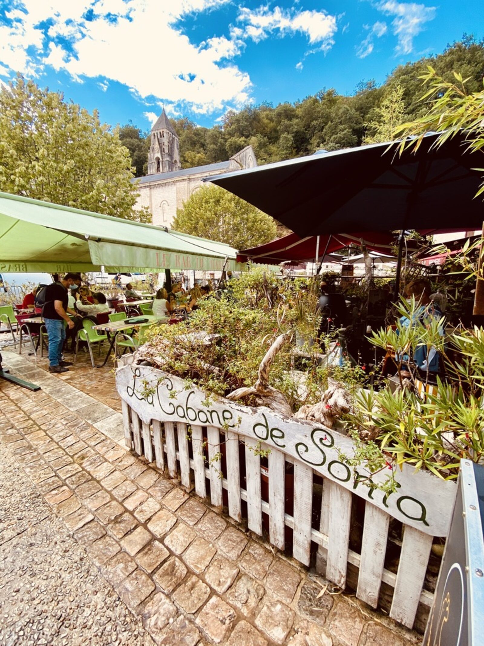 A shaded café terrace in Brantôme — green awnings, black umbrellas, a planter in the foreground with 'La Cabane de Sonia' painted on it, the bell tower of the abbey rising behind through the trees