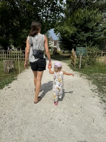 A mother and small child walking hand-in-hand up the gravel drive to Château de Bridoire, the wisteria-shaded approach with the château's pigeonnier visible in the distance through the trees
