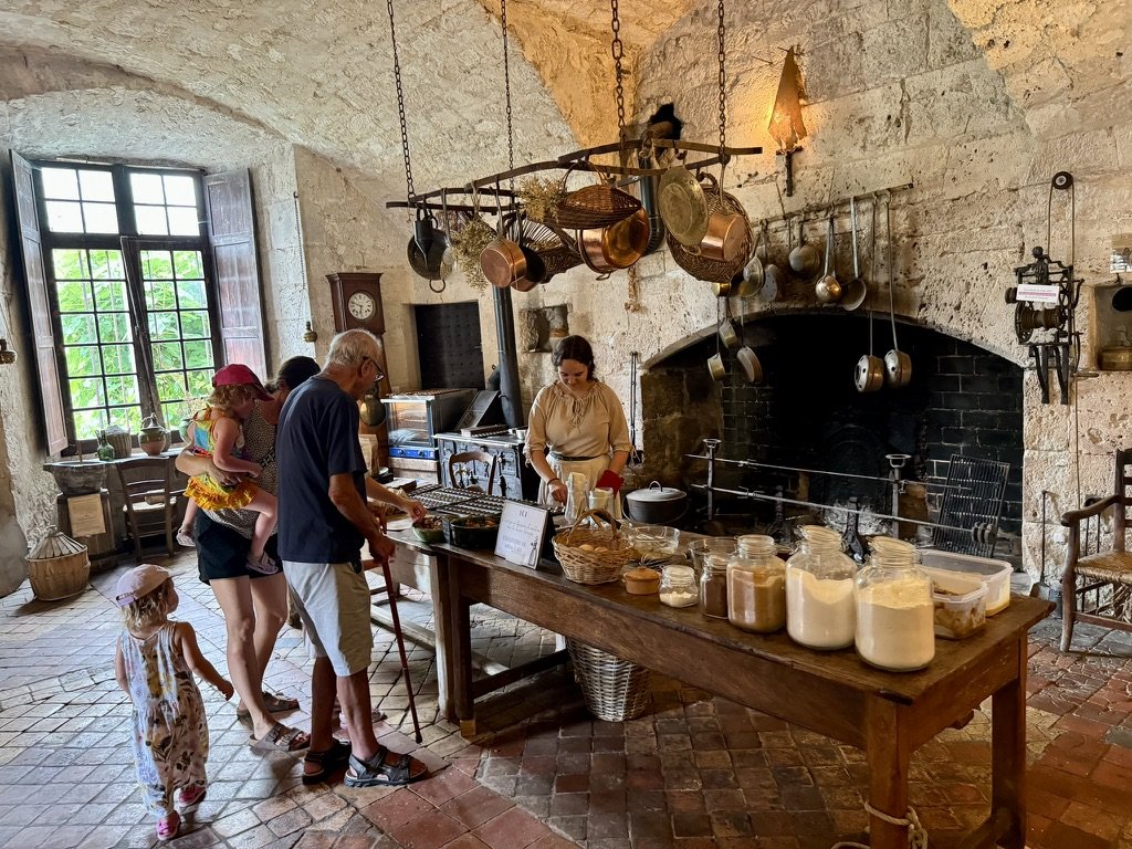 Inside the Bridoire kitchen — stone-vaulted ceiling, copper pans hung from a central wrought-iron rack, blackened fireplace, a costumed staff member behind a long wooden table laid with mason jars of preserved goods