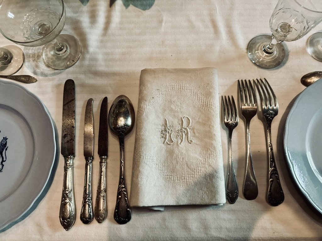 A period table setting at Bridoire — tarnished silver-plate flatware with ornate handles, an embroidered LR-monogrammed linen napkin, antique cut crystal glasses, plates with a heraldic crest, all on a pale linen tablecloth