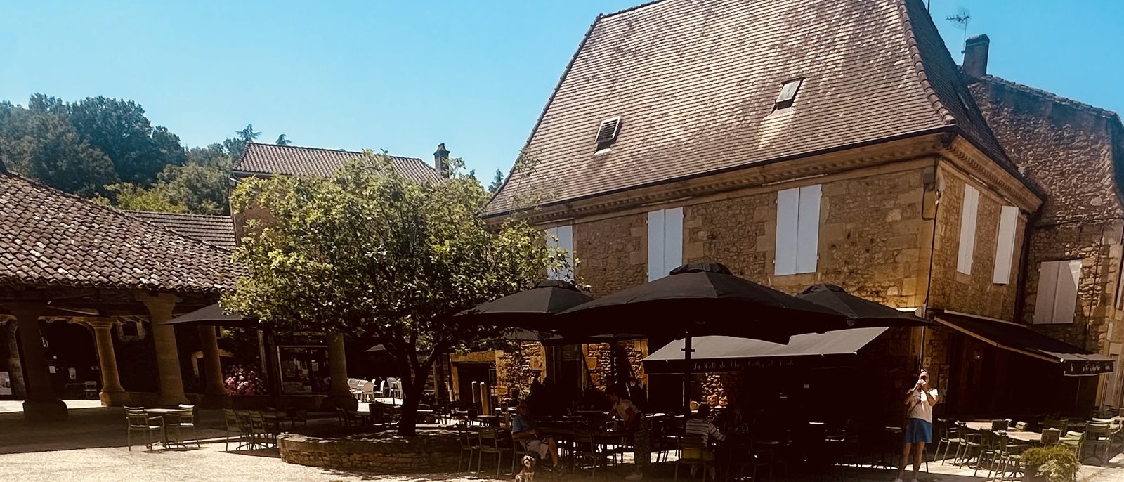 The square under the halles in Cadouin — stone arcades to the left, café terrace under umbrellas, sunlit village houses.