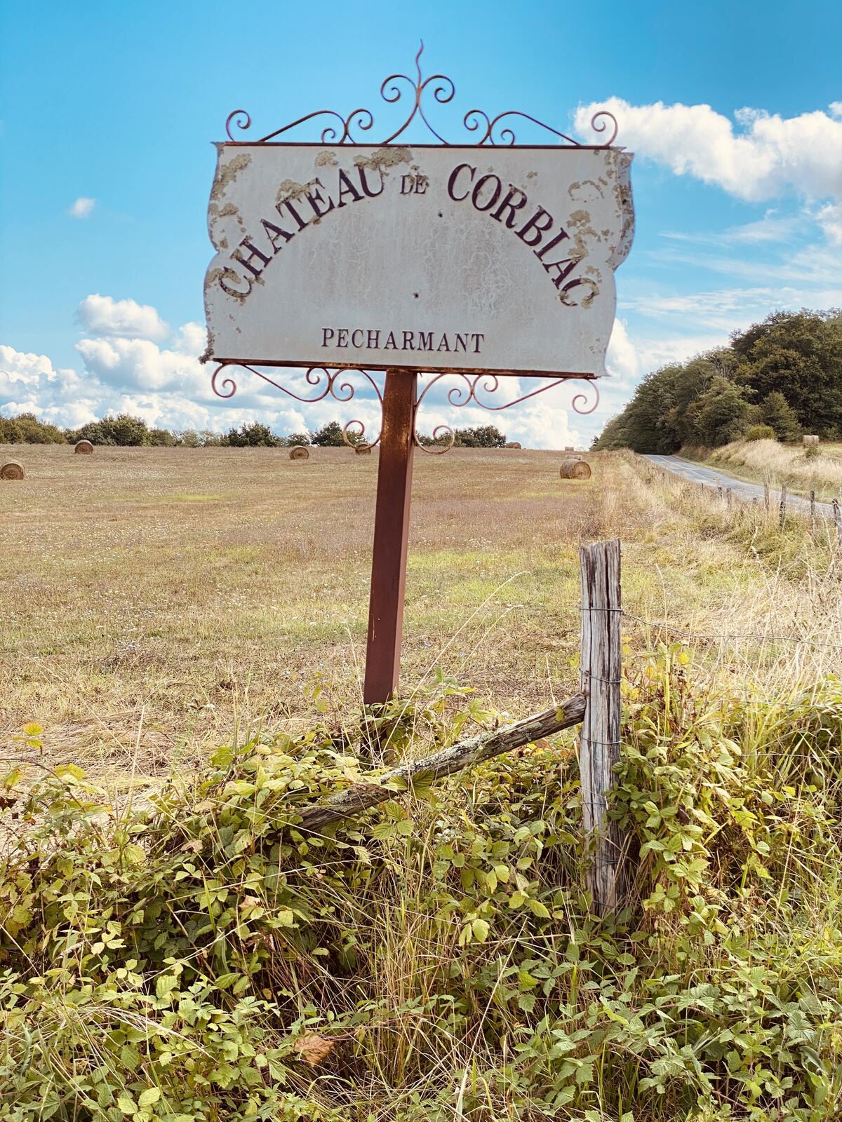 The Château de Corbiac sign standing in a hayfield of round bales — a hand-wrought iron frame with curling scrollwork, a painted metal plate reading CHATEAU DE CORBIAC with PECHARMANT underneath, the paint flaking, a weathered wooden fence post in the foreground and a country D-road running off to the right