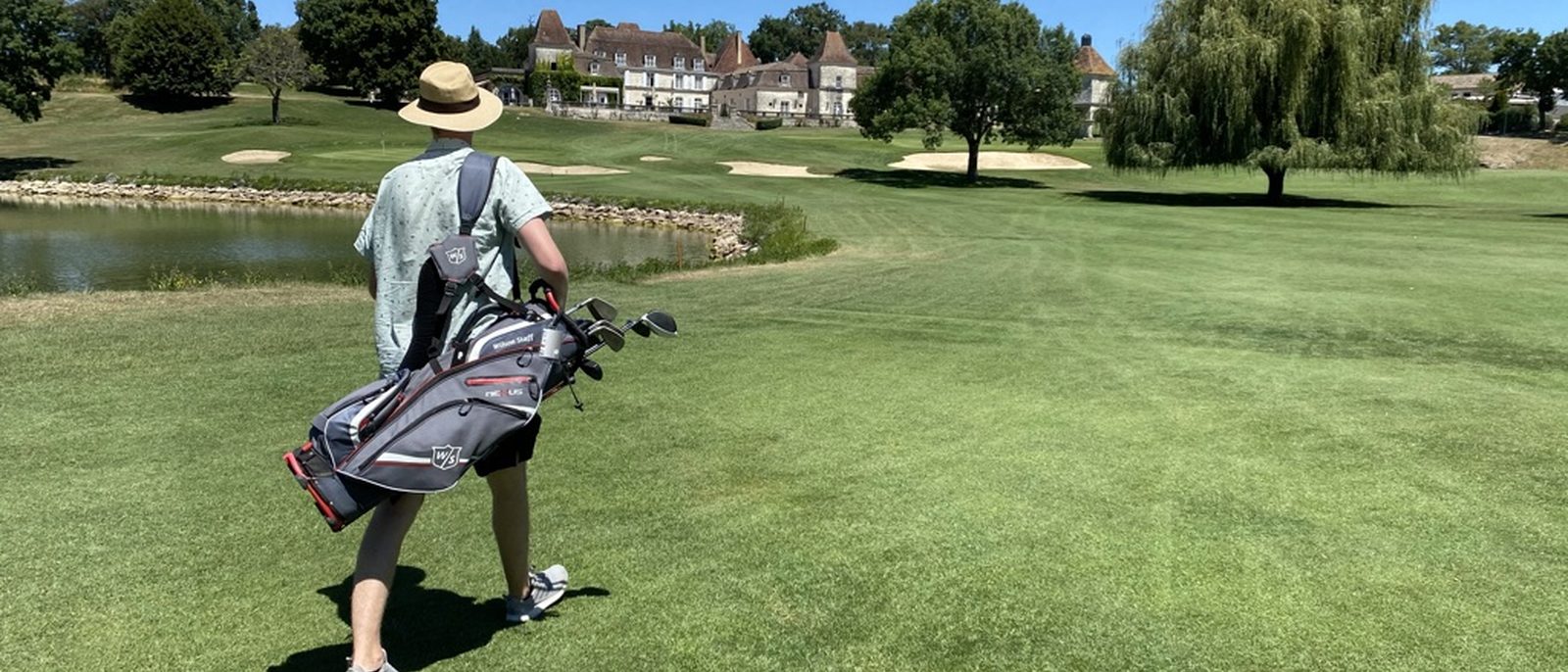 A golfer walking the fairway at Château des Vigiers — the château in the distance, water hazard left, willow tree right.