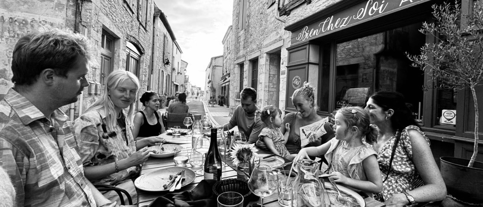 A long lunch on a sun-bright lane in Issigeac — family at a table, sunflower in a vase, wine glasses, the receding street and stone houses framing the scene.