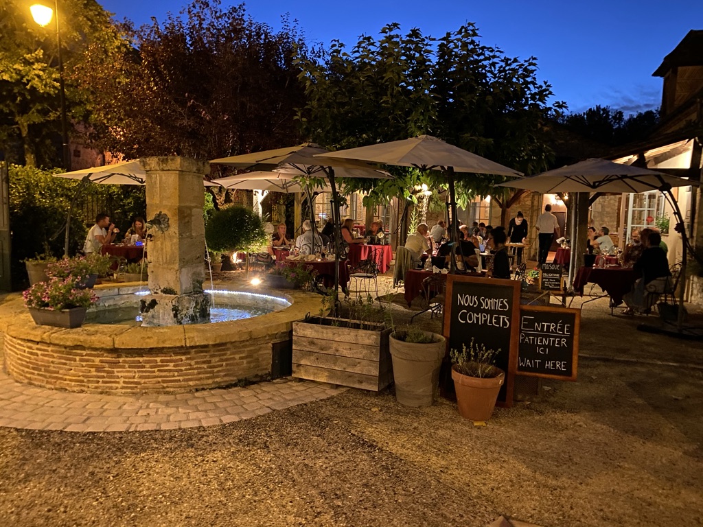 The L'Atelier courtyard in Issigeac at blue hour — stone fountain, white umbrellas, candle-lit tables, a 'Nous sommes complets — Wait Here' sandwich board.