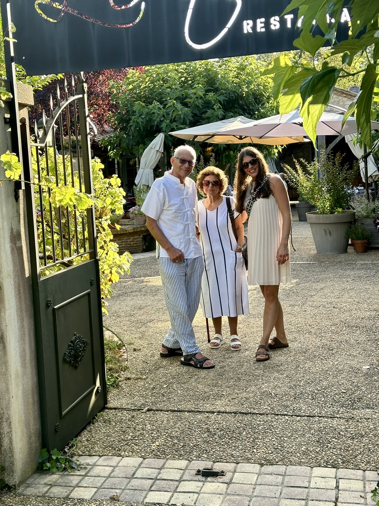 Skip, Stéphanie and Skip's mother at the L'Atelier gate, late afternoon light, the restaurant signage above, courtyard visible behind.