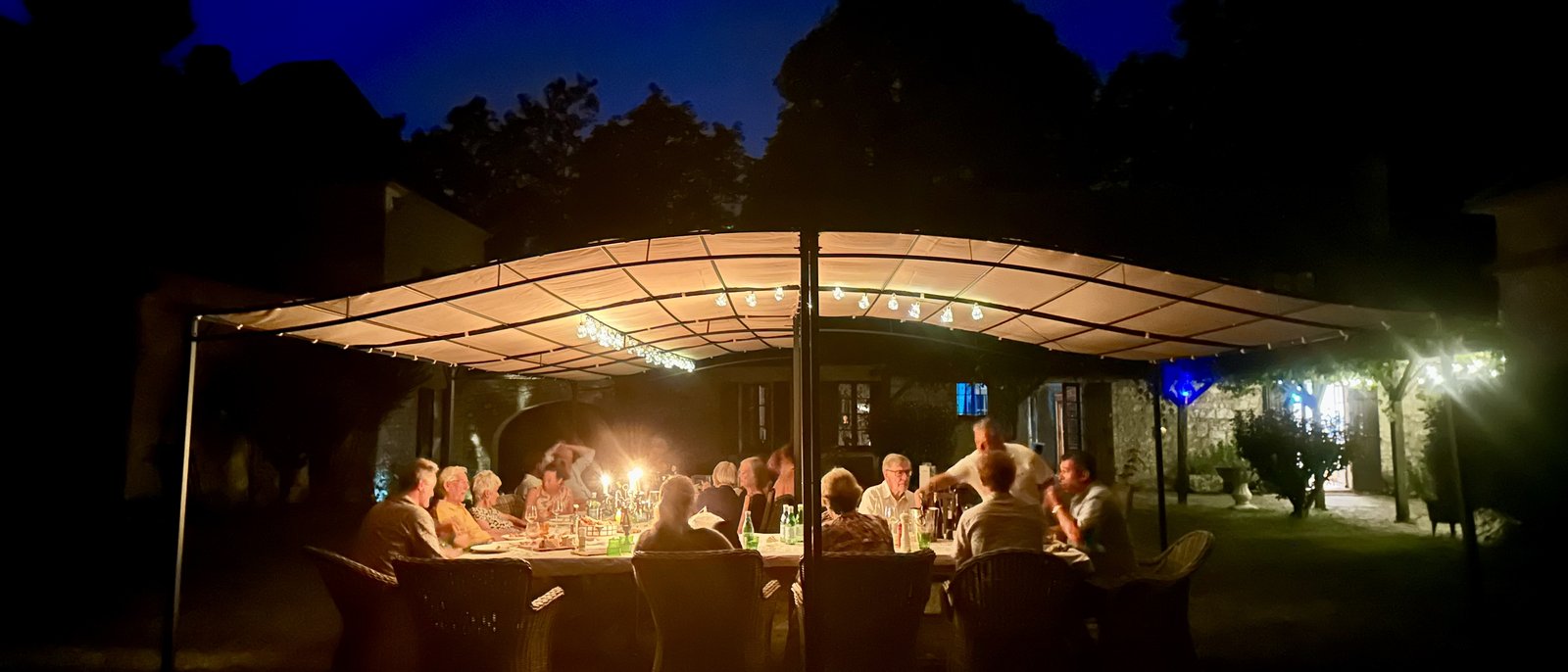 A long dinner under the courtyard pergola at Le Suquet — fairy lights strung overhead, candelabrum centre table, guests on both sides, deep blue summer night sky behind.