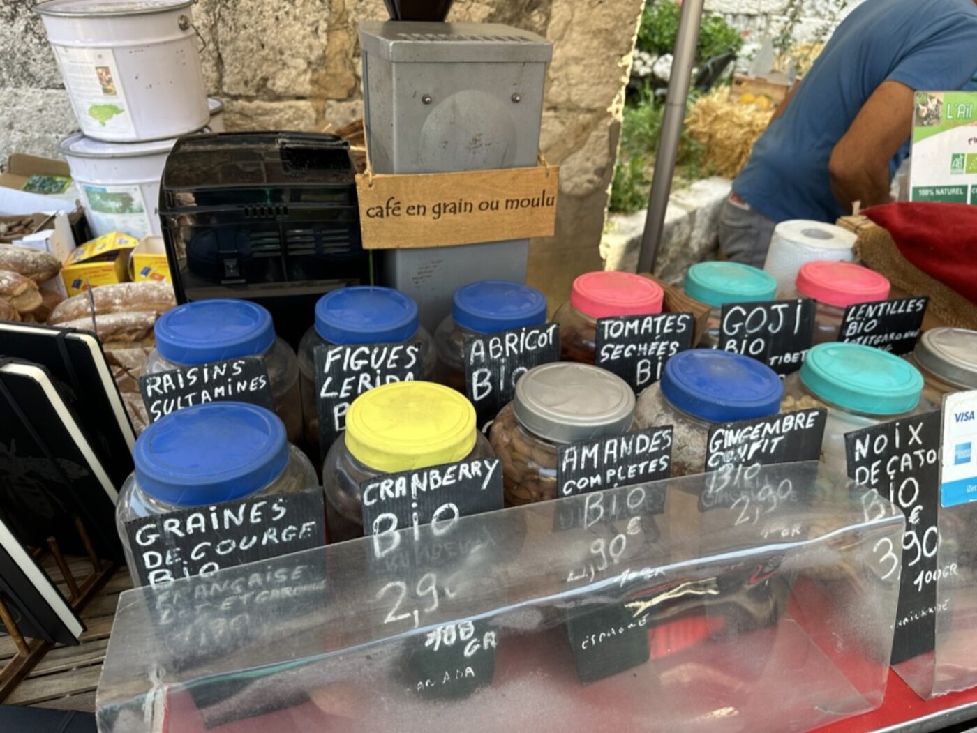 The bio stall at Issigeac market — plastic jars with coloured lids and handwritten chalk labels reading graines de courge bio, raisins sultanines, figues Lerida, abricot bio, tomates séchées bio, goji bio, cranberry bio, amandes complètes, gingembre confit, noix de cajou bio, with a coffee grinder labelled 'café en grain ou moulu'