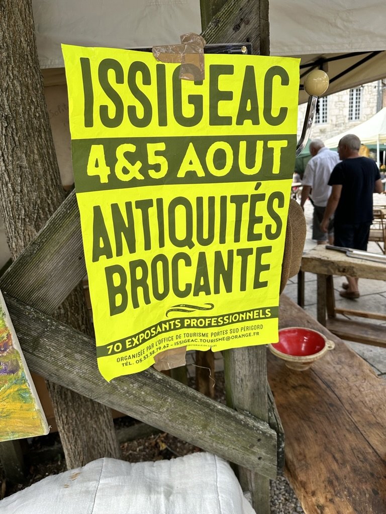 A bright yellow and green poster taped to a wooden post at a brocante stall: ISSIGEAC, 4 & 5 AOÛT, ANTIQUITÉS BROCANTE, 70 EXPOSANTS PROFESSIONNELS — organisée par l'office de tourisme Portes Sud Périgord