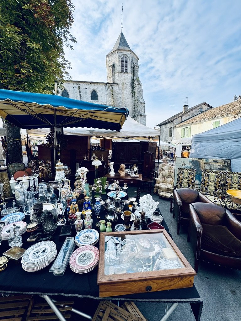 The Issigeac brocante stalls under blue and white striped tarpaulins, with the medieval church spire of Issigeac rising directly behind. Foreground table heaped with brocante material — transferware plates, pewter, glassware, religious figurines, a wooden display case full of silver flatware. To the right, two leather club armchairs and an embroidered ottoman under a French Empire gilt mirror