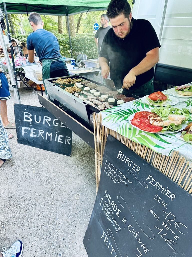 A burger fermier stand at a Périgord summer market — a young chef working a plancha grill, beef patties and rounds of cabécou goat's cheese cooking, plates of cured meats and salad on the counter, a chalkboard menu reading 'Burger fermier seul 12€, avec frites 14€, salade de chèvre, salade-tomate-vinaigrette'.