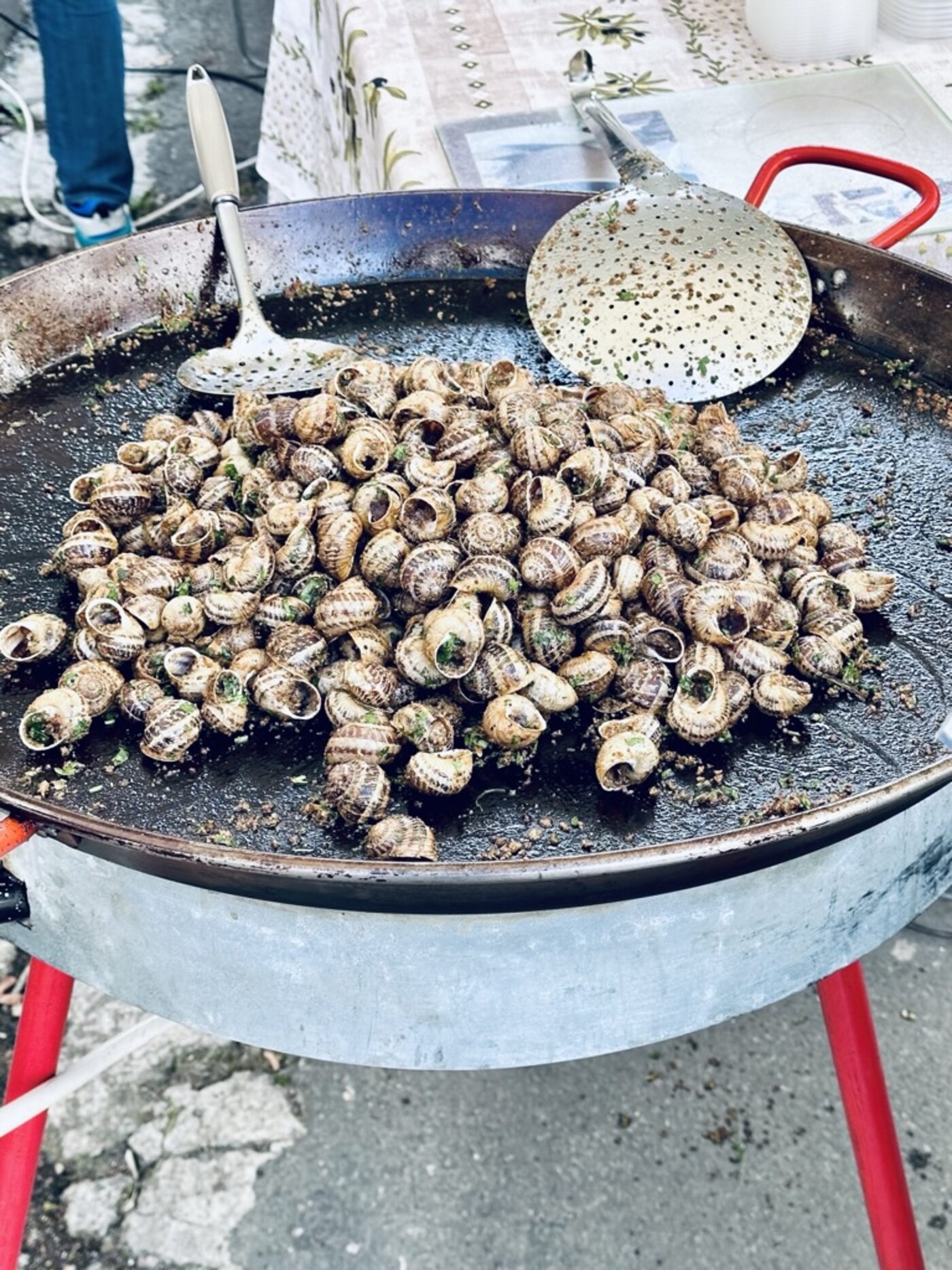 A huge pan of escargots à la plancha — snails cooking in garlic-parsley butter in a large flat steel pan with a slotted skimmer and perforated spoon on top, set up as street food at the Issigeac Sunday market