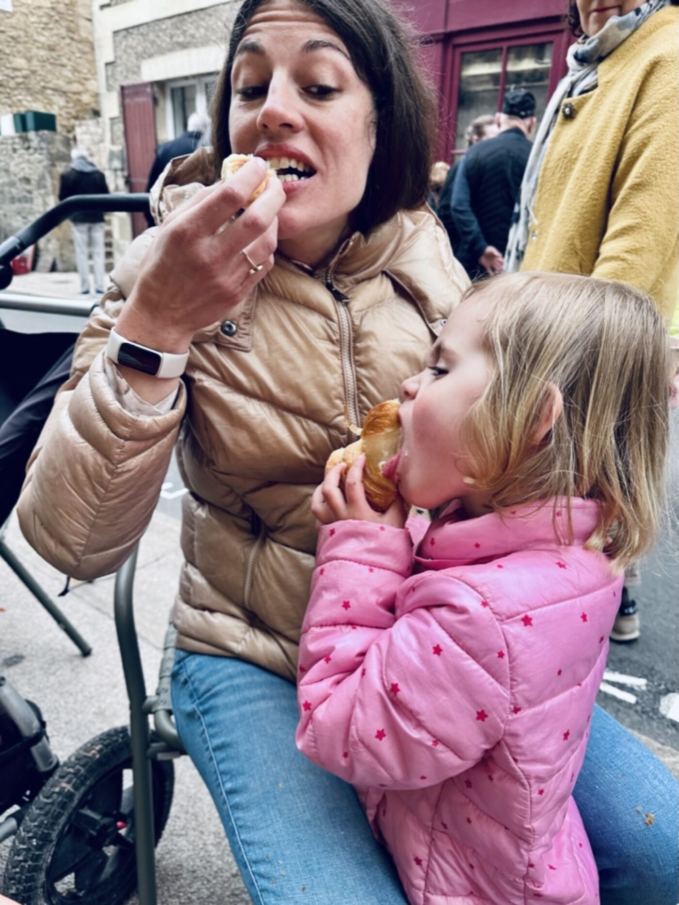 Stéphanie and a small daughter eating pastries at the Issigeac market — Stéphanie in a caramel puffer jacket sharing a croissant with her daughter in a pink puffer jacket with red stars, outdoor market scene behind, autumn light