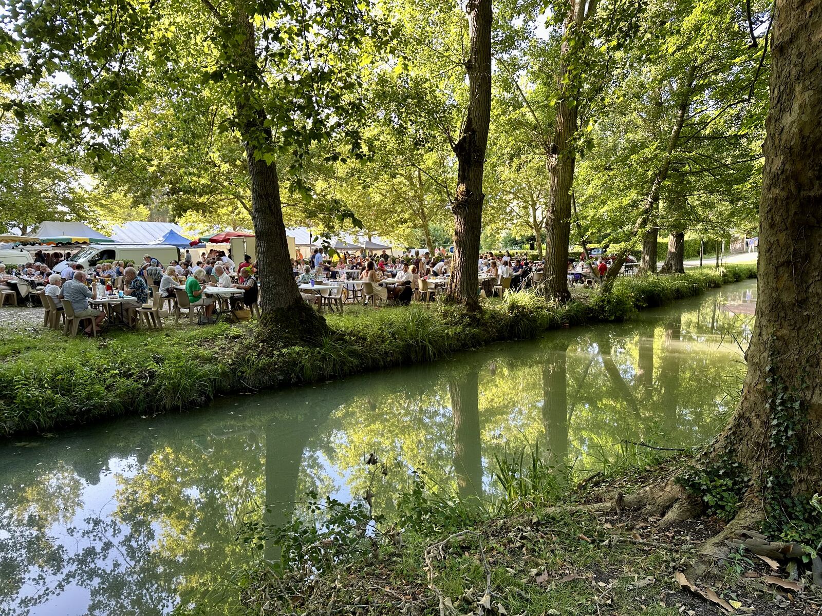 The Issigeac Thursday night market in summer — hundreds of people seated at long trestle tables under the dappled shade of tall plane trees along a narrow tree-lined stream, food vans and tents in the background, families and older couples sharing dinner in the soft evening light, the stream reflecting the leaves