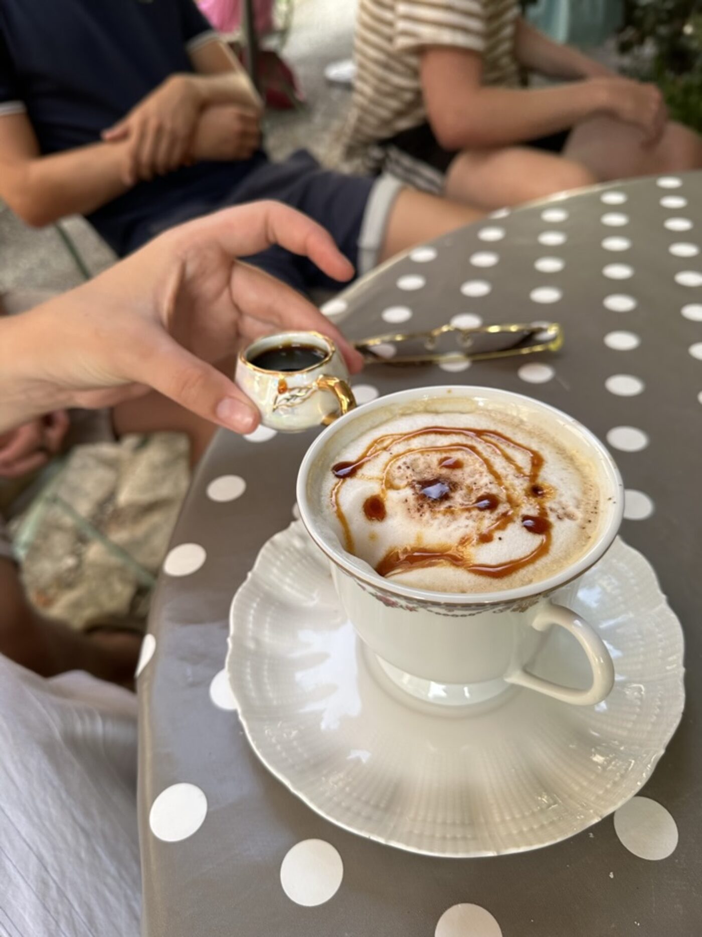 A cappuccino on a polka-dot enamel table at Shabby Chic — cream foam with a caramel drizzle swirl on top, served on a gold-rimmed porcelain saucer, with a tiny matching porcelain cup of espresso alongside held by a hand, Sunday morning market scene visible in the background