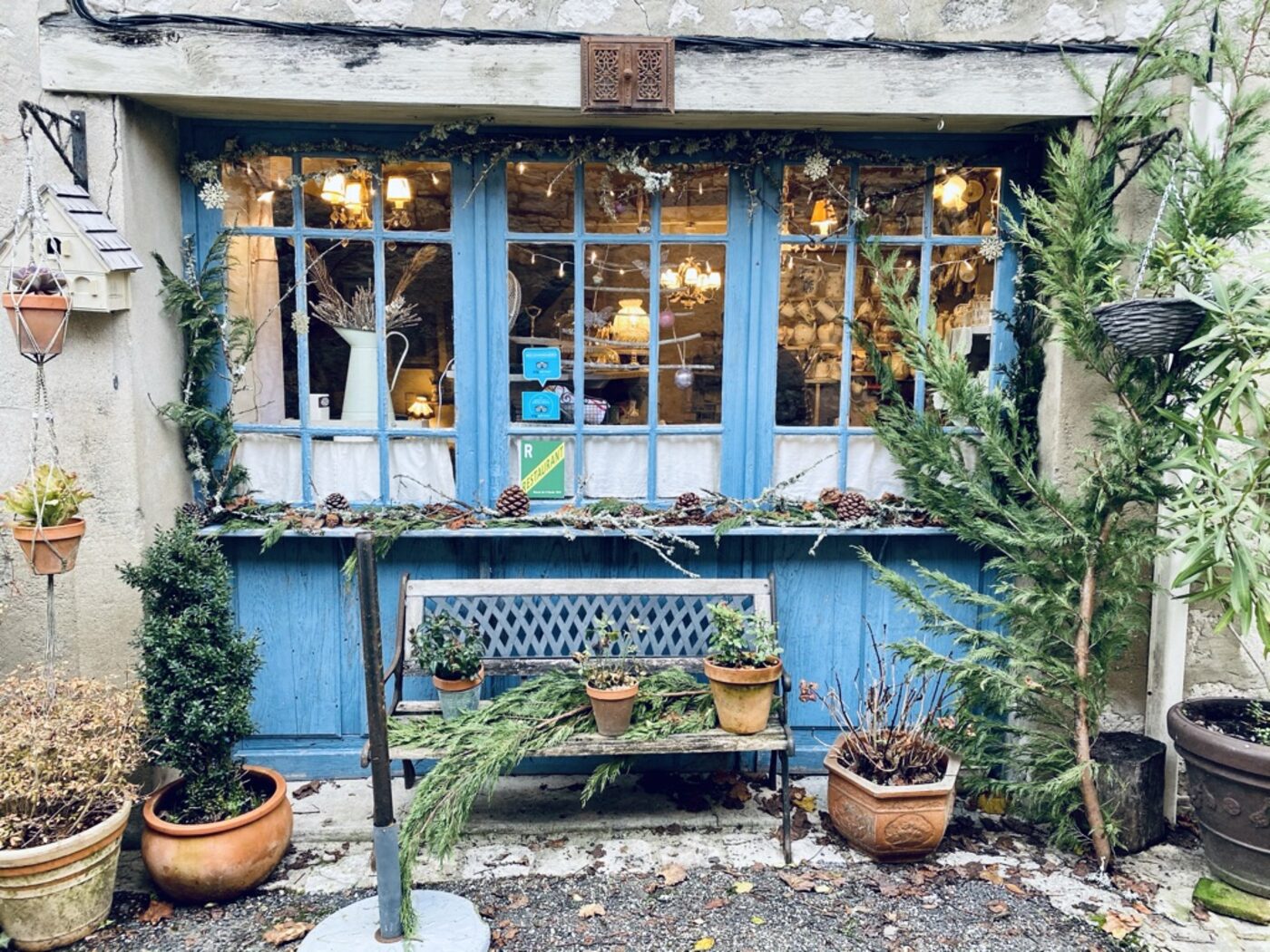 The facade of Shabby Chic café in Issigeac — a blue-painted bifold-door wooden shop-front with warm lights inside, antique lamps and chandeliers visible through the glass, a weathered bench in front with potted plants and conifer branches, winter light