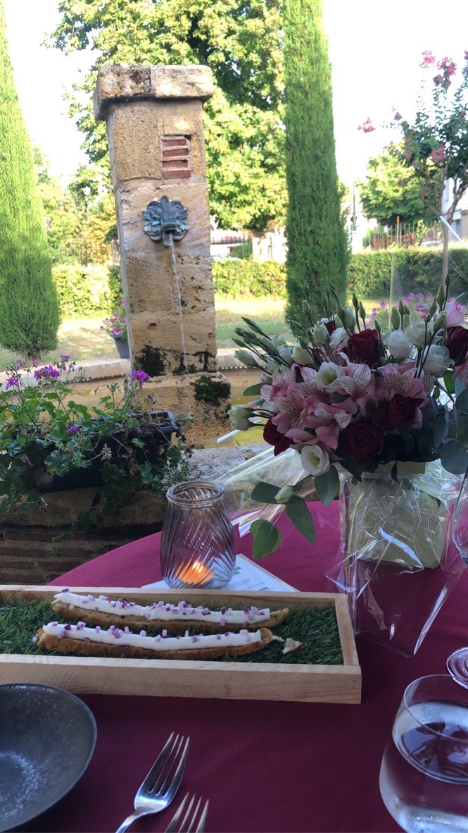 A garden dining table at Bignac with a stone fountain behind, an asparagus amuse-bouche on a wooden tray, fresh flowers, candle, late-afternoon light