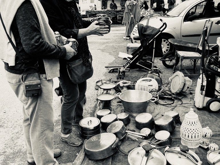 Black-and-white photo of Pat and Geoff at a country brocante, examining cookware spread on a tarpaulin