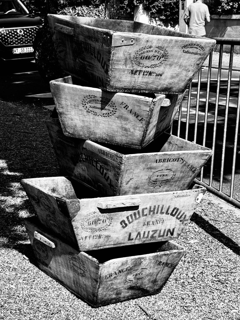 A black-and-white photo of stacked French wooden fruit crates with original stencil printing visible: BOUCHILLOUX LAUZUN, FRANCE, ABRICOTS, PRUNEAUX D'ENTE 60/70, NET 12K500. Five crates piled into a rough pyramid on a sunny pavement