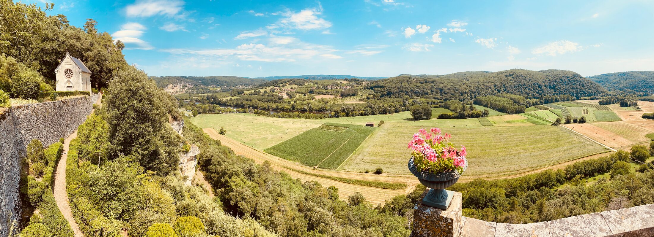 A panoramic view from the Marqueyssac belvedere — a small stone chapel on the left, a cast-iron urn of pink geraniums in the foreground, and the whole Dordogne valley unfolding below with river, fields, villages, and hills