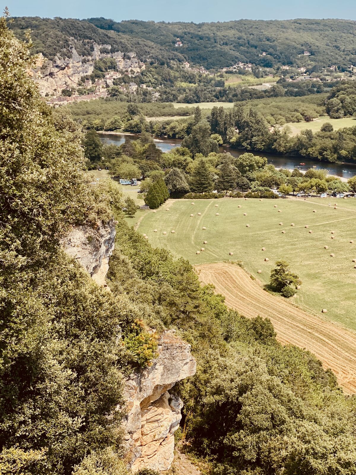 The view down from Marqueyssac — limestone cliffs on the left, La Roque-Gageac village clinging to them, the Dordogne river curving through hayfields below with small canoes on the water
