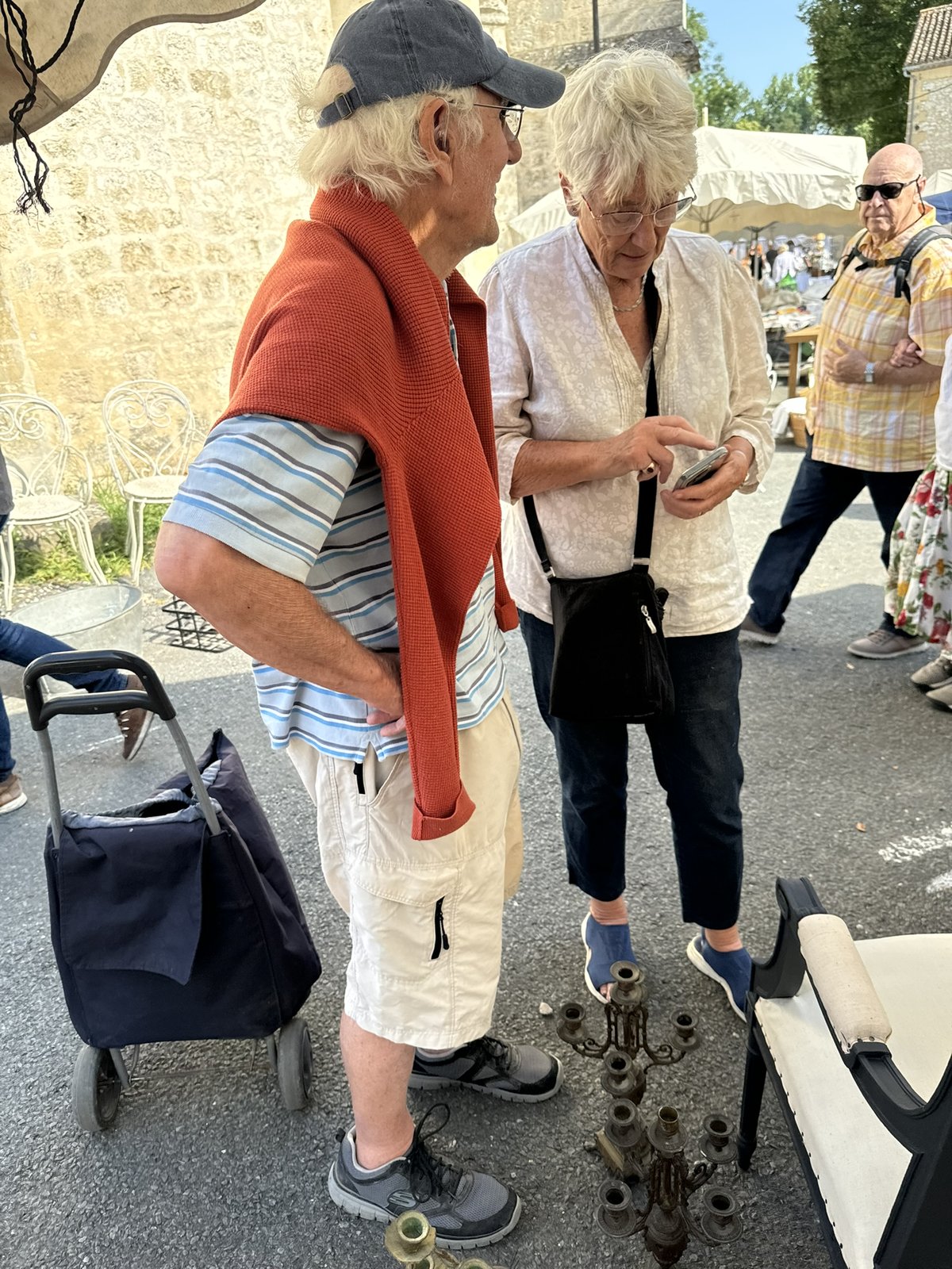Geoff and Pat at a brocante, mid-decision. Geoff in a cap and orange jumper tied across his shoulders, holding the wheeled shopping bag for transporting purchases, watching Pat as she examines something on her phone. A multi-arm brass candelabrum on the ground in front of them, partly out of frame