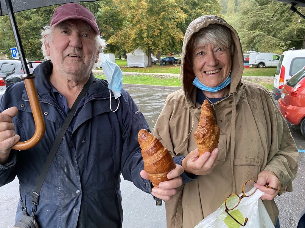 Pat and Geoff laughing in the rain at a brocante, each holding a buttery pain-au-chocolat. Geoff in a red baseball cap holding an umbrella, mask pulled below his chin; Pat in a beige hooded raincoat, glasses in her free hand, both grinning at the camera