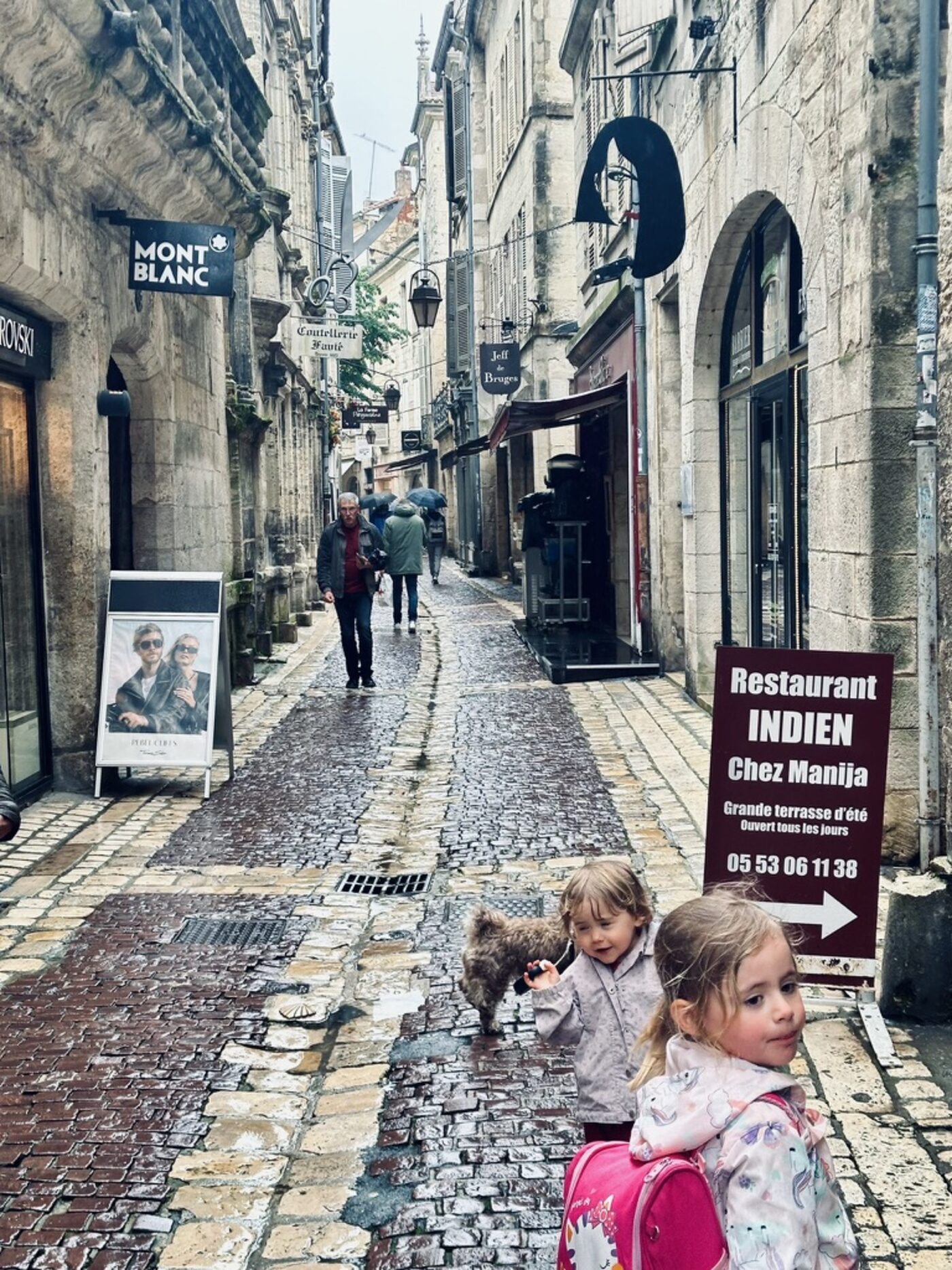 A cobbled street in the Périgueux old town on a rainy day — two small girls in the foreground with a small dog, mediaeval stone houses with iron signs, a dark-red sign on the right reading 'Restaurant INDIEN — Chez Manija'
