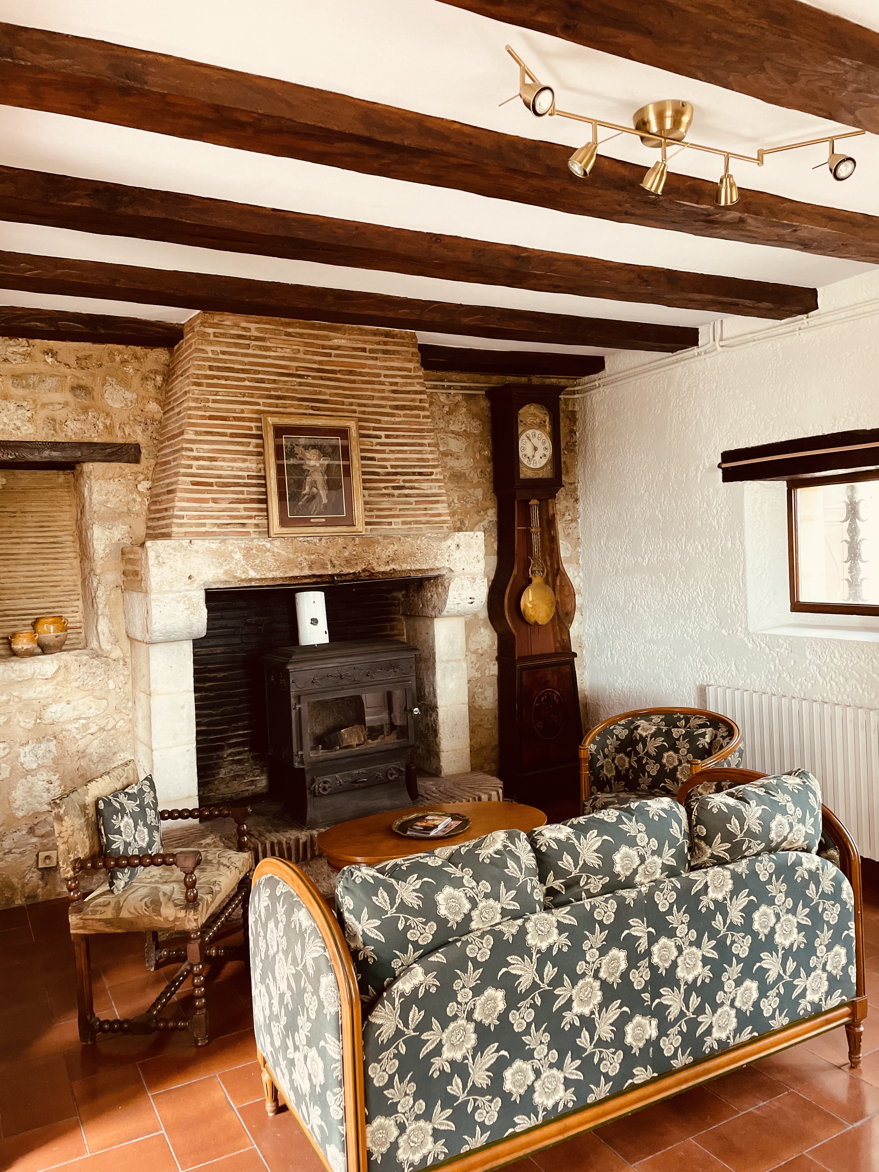 The salon at Le Pigeonnier, with a wood burner set into a brick and stone hearth, a Périgord longcase clock and a green floral sofa