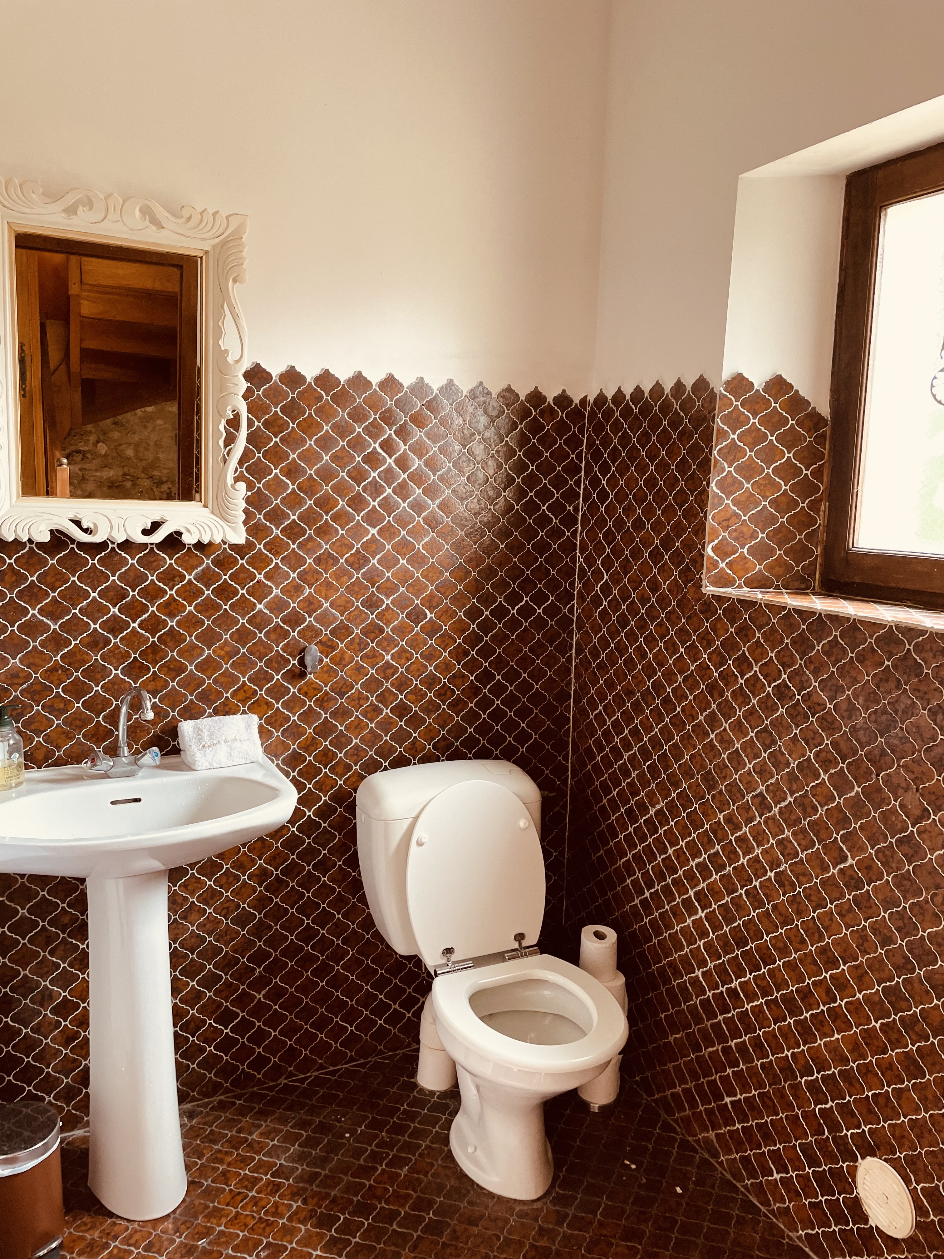 The first floor shower room at Le Pigeonnier, tiled to shoulder height in brown arabesque zellige, with a pedestal basin and a carved white mirror