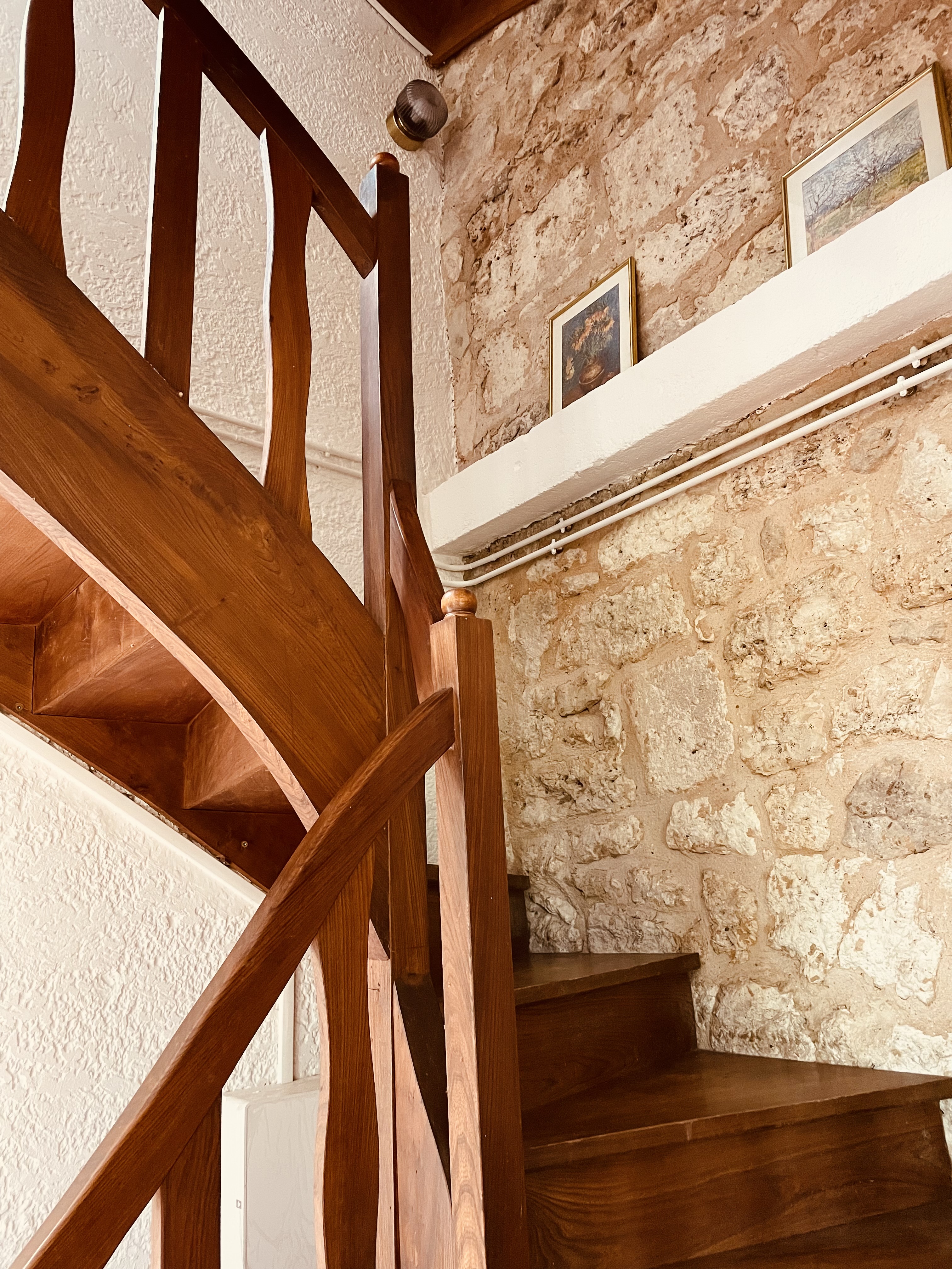 The timber staircase at Le Pigeonnier turning against a bare stone wall hung with framed watercolours