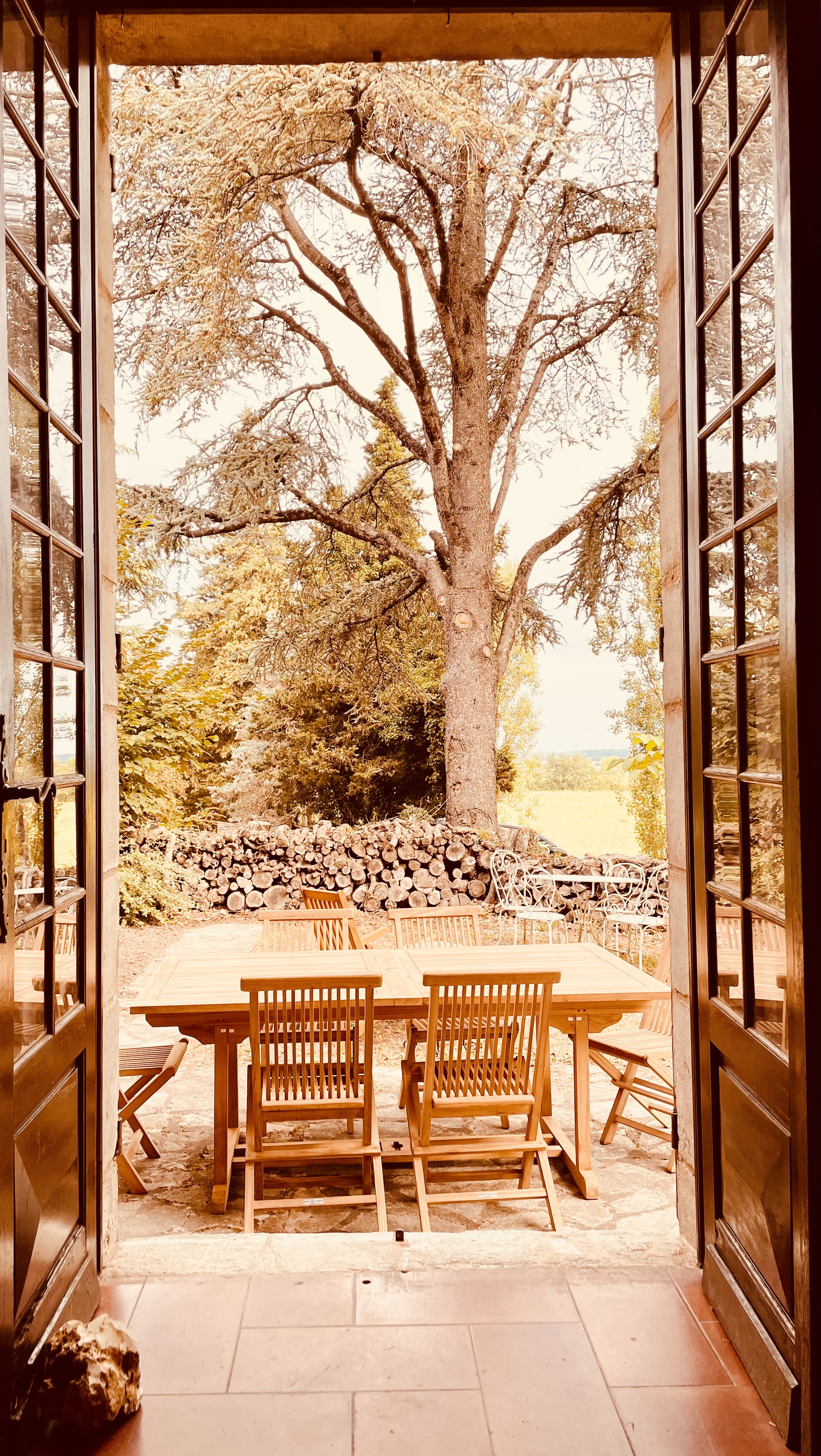 The terrace at Le Pigeonnier seen through open French doors, with a teak table under a large cedar and a wall of stacked firewood behind