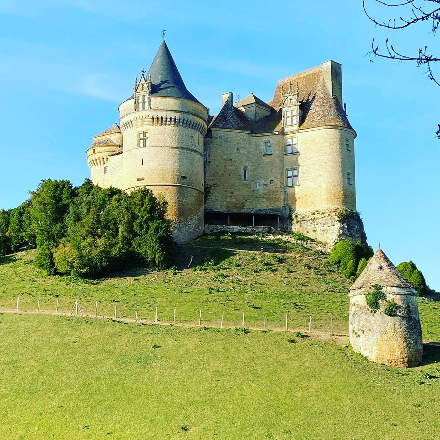 Château de Bannes at golden hour, perched on its hilltop with the conical-roofed corner tower catching low sun, machicolated walls visible, and a small stone pigeonnier on its own knoll in the foreground