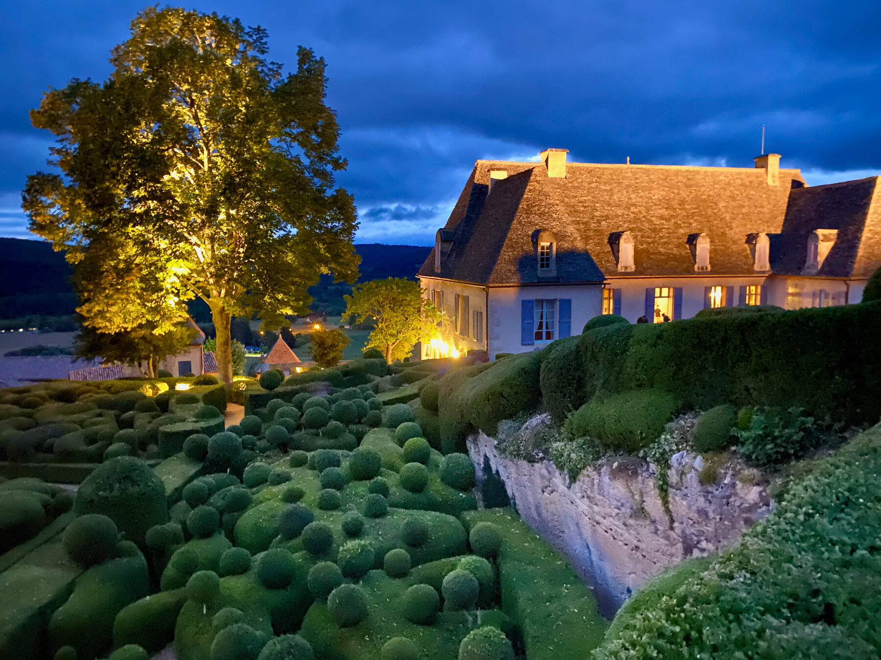 Les Jardins de Marqueyssac at blue hour — the clipped boxwood topiary lit from below, the chateau glowing yellow against a dusk sky, the cliff edge above the Dordogne valley