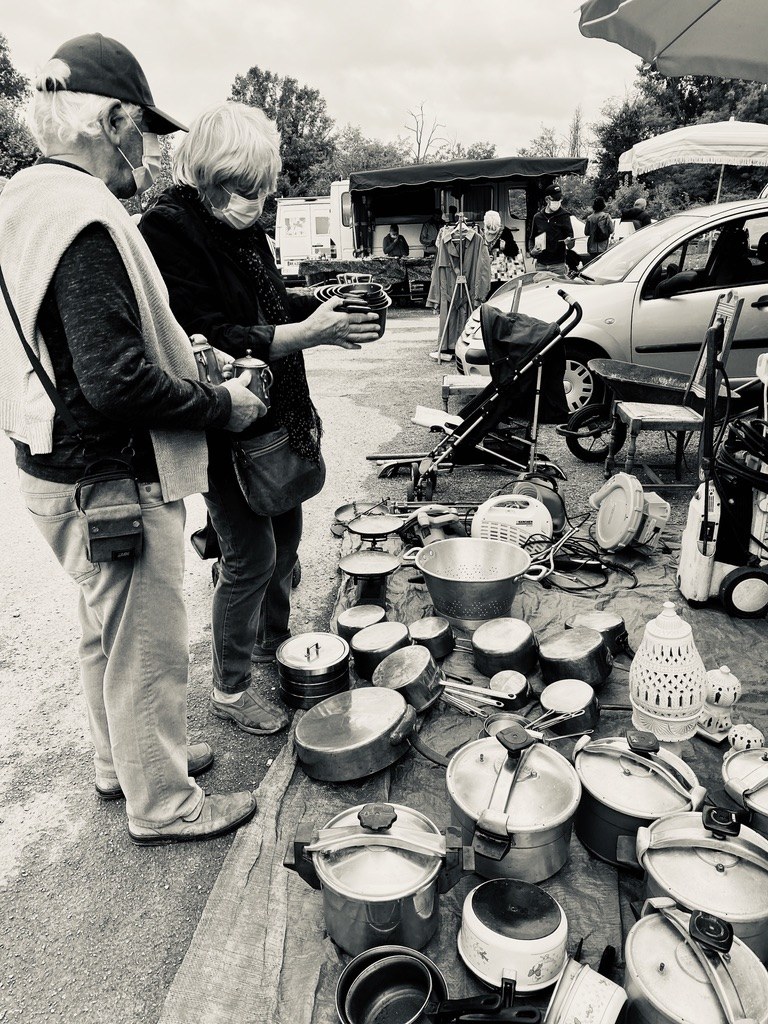 Black-and-white photo of Pat and Geoff at a country brocante, examining a piece of enamelware. Geoff in profile in a cap with a sleeveless cardigan tied over his shoulders; Pat next to him with white hair, holding the enamelware carefully. A tarpaulin spread before them with cast-iron pans, pressure cookers, kitchen scales, an enamel colander, and lanterns