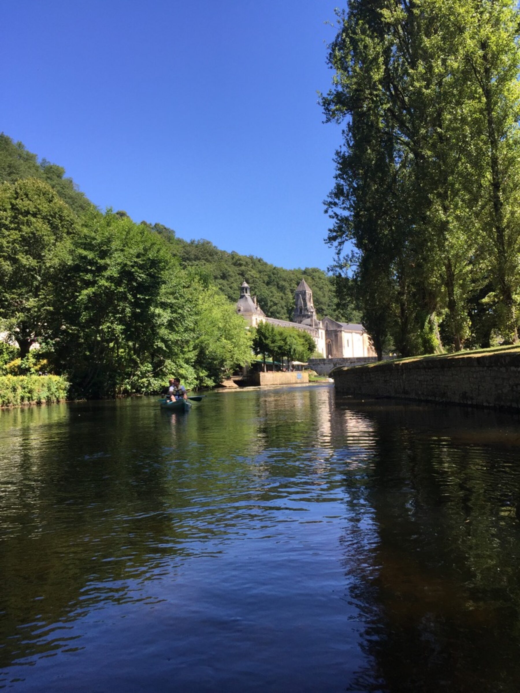 A canoe on the Dronne river approaching the abbey of Brantôme — summer, deep reflections, the abbey's distinctive bell tower in the distance between the trees