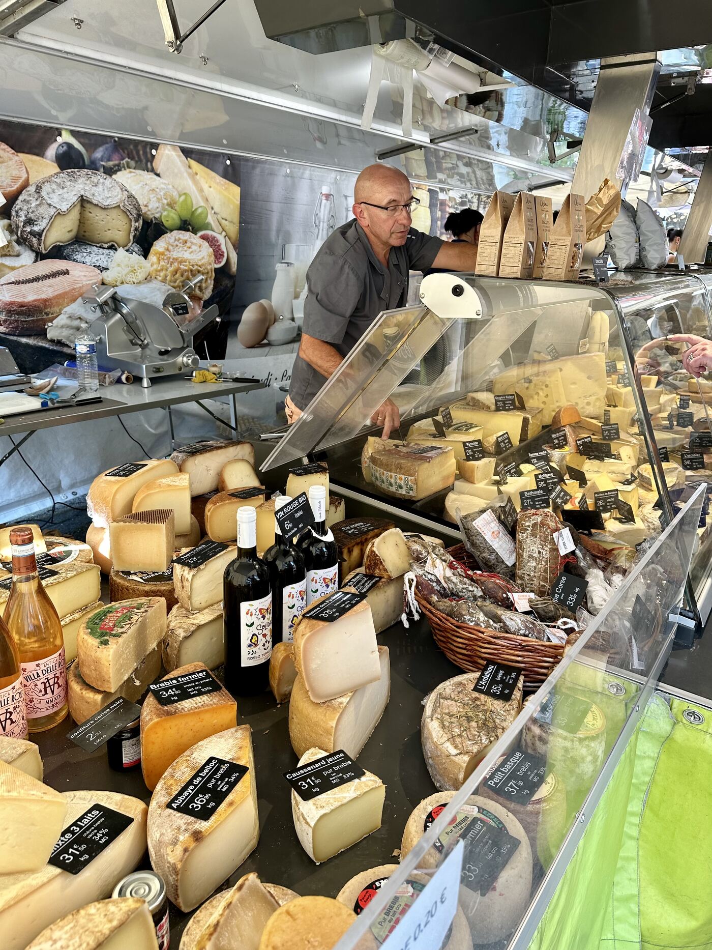 The cheese and charcuterie van at the Saint-Astier Thursday market — a serious fromager leaning over his case of hard cheeses, wheels of Abbaye de Belloc, Caussenard, Petit Basque, Ardalhou, Pecorino, Gruyère, Bethmale and pyrenean sheep's milk cheeses stacked in front, a basket of Corsican salami beside him, bottles of natural wine tucked between the wheels, prices chalked at 31 to 37 euros a kilo