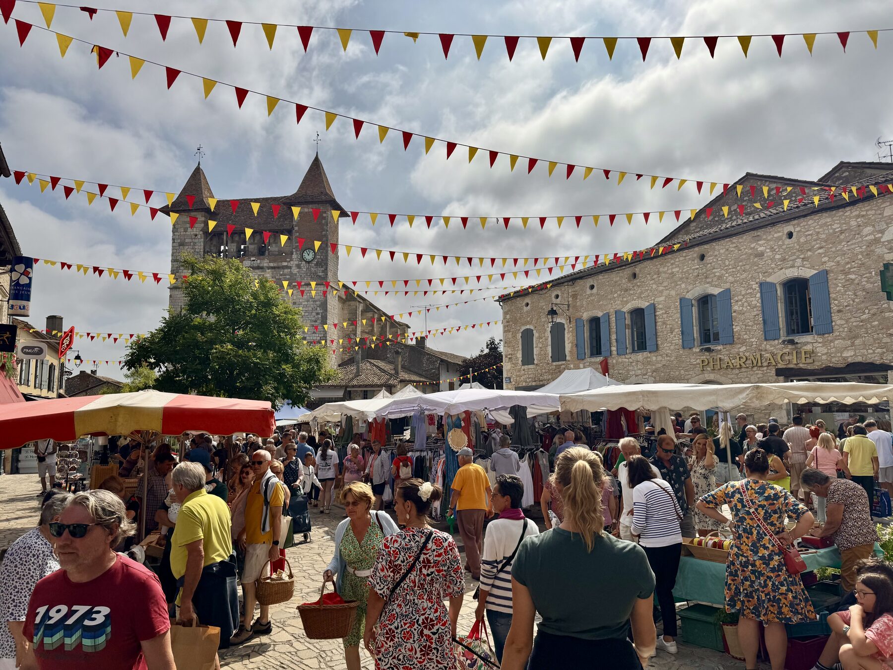 Villeréal Saturday market in summer — the medieval bastide tower of the fortified église Notre-Dame rising above warm-stone houses, red-and-yellow bunting strung across the square overhead, dozens of market stalls with white tarpaulins and tents, crowds of shoppers in summer clothing wandering between the stalls, a Pharmacie sign on a golden-stone building on the right