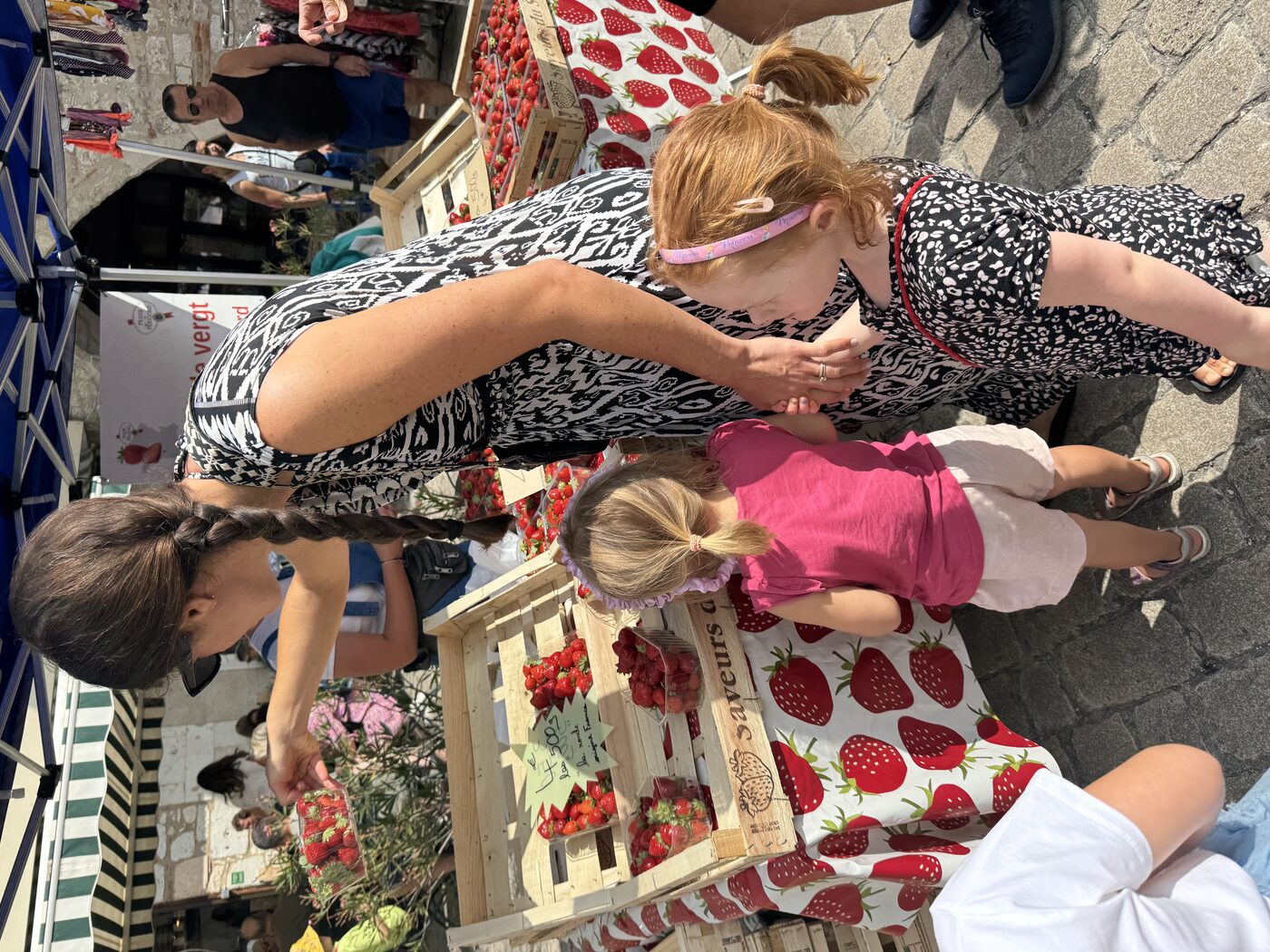 Stéphanie and our two daughters at a strawberry stall at Villeréal Saturday market — a wooden crate stacked with punnets of red strawberries labelled 4€ les 500g, Saveurs du Périgord branding on the crates, a Fraises du Périgord IGP rosette poster above