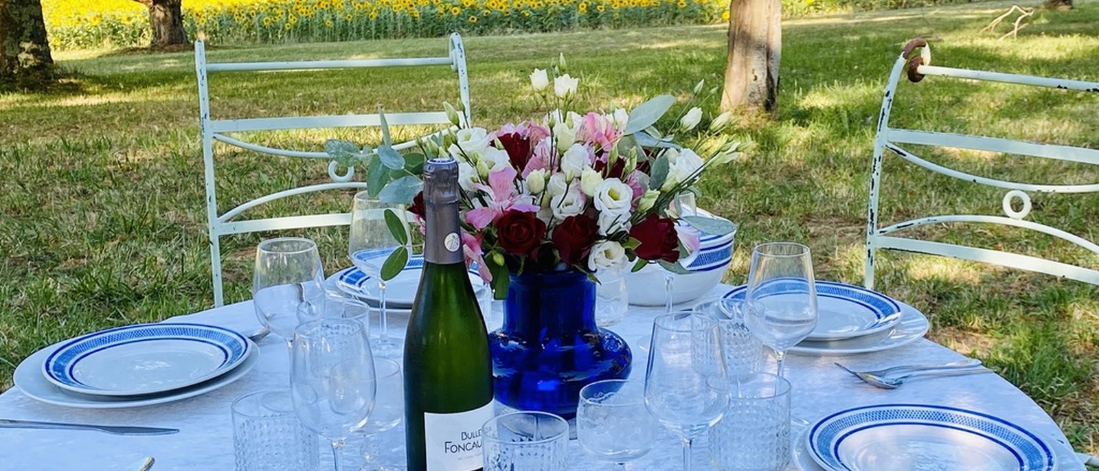 A summer table at Manoir du Suquet, set under the trees with a sunflower field beyond — blue-and-white Limoges plates, crystal glasses, a bottle of Pét-Nat at centre.